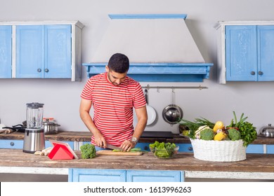 Young Man In Casual T-shirt Chopping Cutting Vegetables, Preparing Vegetarian Salad In Modern Kitchen, Cooking Healthy Breakfast, Basket Of Fresh Green Vegetables On Table, Vegan Food, Diet Nutrition