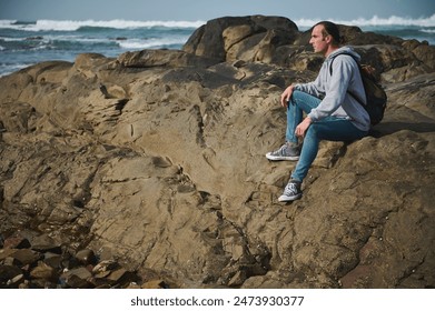 Young man in casual wear sitting on rocky terrain along the coastline. The ocean waves crashing in the background add to the serene outdoor atmosphere, emphasizing adventure, travel, and tranquility. - Powered by Shutterstock