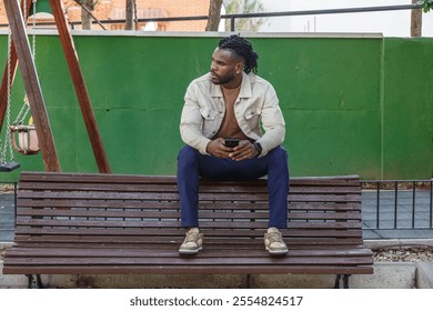 A young man casually dressed in a light jacket and dark pants sits alone on a wooden bench. He is holding a smartphone and looking contemplatively into the distance, surrounded by a green park. - Powered by Shutterstock