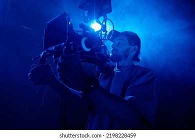 Young Man In Casualwear Holding Video Camera While Shooting Commercial In Studio Lit With Blue Light And Full Of Smoke