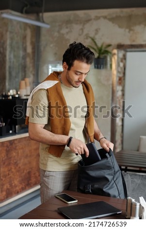 Young man in casualwear standing by table with mobile gadgets and putting notebook into black bag while leaving cafe after having lunch