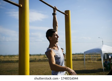 A Young Man Of Caucasian Appearance Pulls Himself Up On A Horizontal Bar. The Guy Is Hanging On One Arm. Healthy Lifestyle, Affordable Outdoor Sports