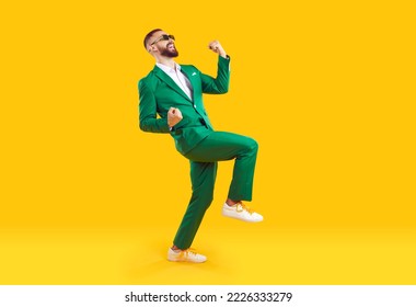 Young man celebrating success. Happy funny joyful excited guy in stylish green party suit and cool glasses raising fist up and dancing isolated on bright yellow background. Full length shot, side view - Powered by Shutterstock