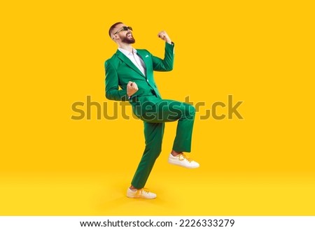 Young man celebrating success. Happy funny joyful excited guy in stylish green party suit and cool glasses raising fist up and dancing isolated on bright yellow background. Full length shot, side view