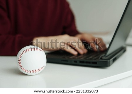 Young man checking baseball stats or writing baseball article on laptop. White leather ball on the table. Betting, gambling concept.