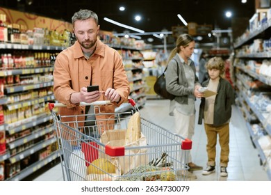 Young man checking phone while pushing shopping cart in grocery store with woman and child in background examining products on shelf