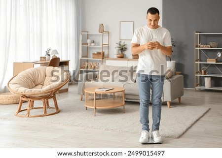 Young man checking weight on digital scales while using smartphone in modern living room, health and fitness monitoring