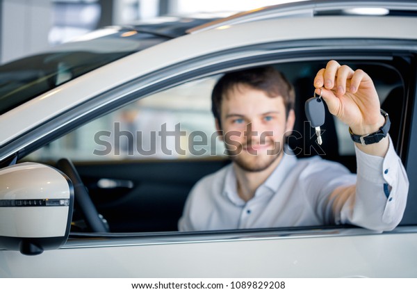 Young man cheering and holding key sitting in\
his new car in showroom