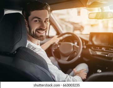 Young Man Is Choosing A New Vehicle In Car Dealership. Looking At The Camera And Smiling.