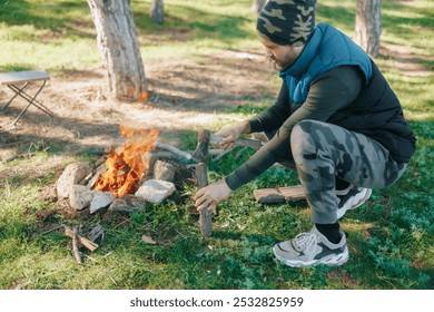 A young man is chopping wood with an axe near a blazing campfire, set in the wilderness with green grass and nature around him, showcasing essential survival skills and campfire preparation. - Powered by Shutterstock