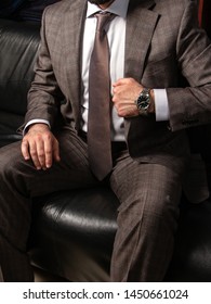 A Young Man In A Classic Suit And Expensive Wristwatch Sits In A Tense Pose On A Black Leather Sofa.  Demonstration Of Excellence. Menswear Italian Designers For The Solemn Occ