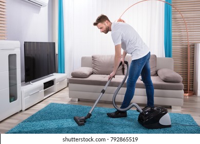 Young Man Cleaning Blue Carpet With Vacuum Cleaner At Home