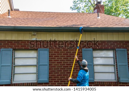 Young man cleaning the soffit of a house with a brush on a long pole. The house has blue shutters and is one story high
