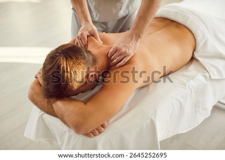 Young man client lying on massage table receiving remedial body back and shoulder massage from professional masseur or doctor physiotherapist in clinic. Physiotherapy and rehabilitation concept.