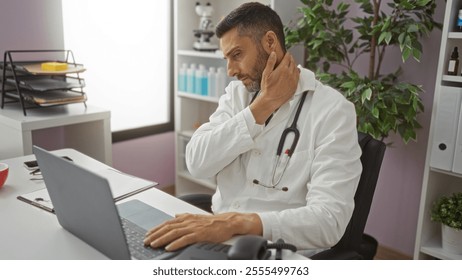 Young man in clinic wearing white coat with stethoscope sits at desk, rubbing neck in discomfort, conveying a busy healthcare professional setting. - Powered by Shutterstock