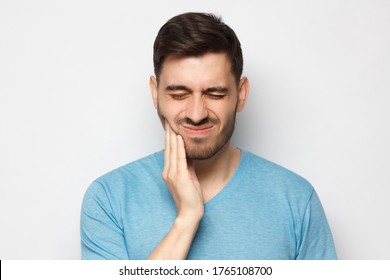 Young Man With Closed Eyes Suffering From Severe Toothache, Touching Jaw With Fingers Trying To Ease Strong Tooth Pain, Isolated On Gray Background