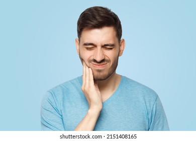 Young Man With Closed Eyes Suffering From Severe Toothache, Touching Jaw With Fingers Trying To Ease Strong Tooth Pain, Isolated On Blue Background