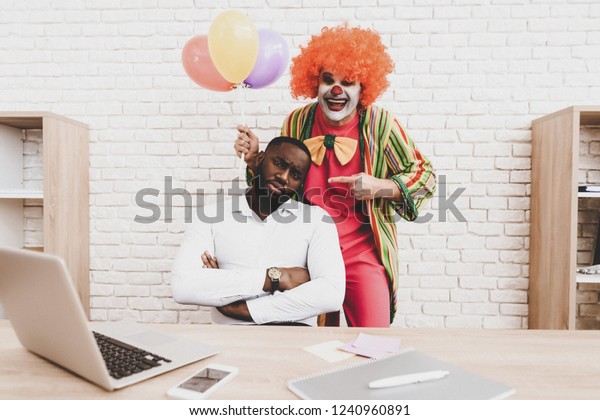 Young Man in Clown Costume with Baloons in Office.\
April Fools Day. Businessman in Office. April Jokes. Workers on\
Meeting. Holidays and Celebration Concept. Clown with Baloons. Man\
with Red Nose.