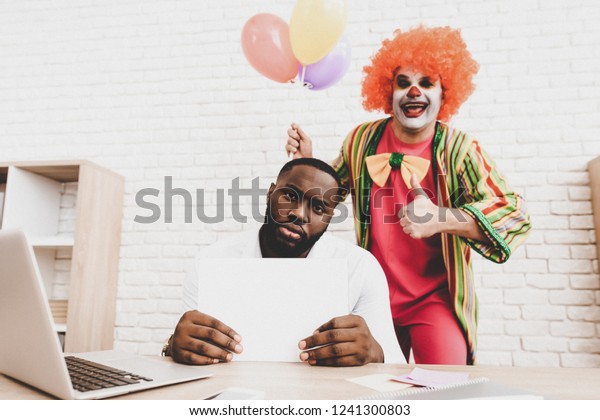 Young Man in Clown Costume with Baloons in Office.\
April Fools Day. Businessman in Office. April Jokes. Workers on\
Meeting. Holidays and Celebration Concept. Clown with Baloons. Man\
with Red Nose.