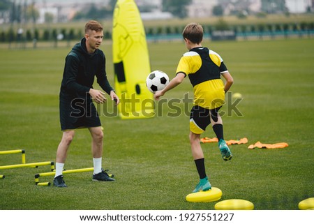 Young Man Coaching Soccer Boy. Coach on Training Session With Teenage Boy. Soccer Player Kicking Ball Standing on Stability Cushion on Grass Field. Football Player Improving Skills With Trainer