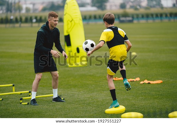 Young Man Coaching Soccer Boy. Coach on Training\
Session With Teenage Boy. Soccer Player Kicking Ball Standing on\
Stability Cushion on Grass Field. Football Player Improving Skills\
With Trainer