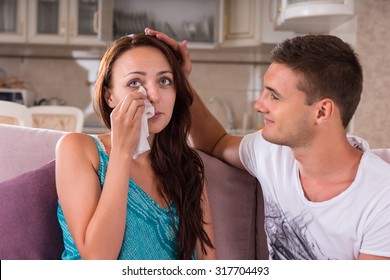 Young Man Comforting Crying Woman Dabbing Her Eyes With Tissue While He Strokes Her Hair And Smiles - Young Couple Watching Emotional Television Program Or Film Together At Home