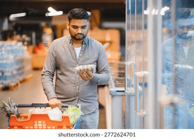 Young man compares prices while holding an egg carton and pushing a full shopping cart down a supermarket aisle, choosing groceries