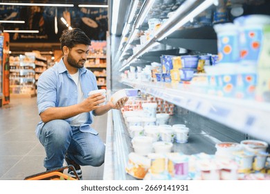 Young man comparing yogurts while crouching in the refrigerated dairy aisle of a grocery store