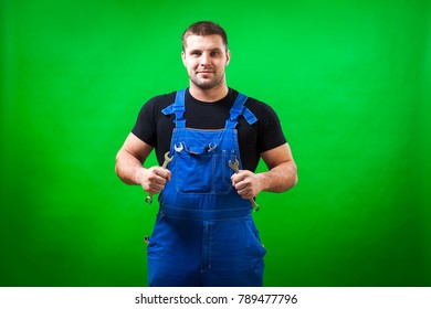 A young man construction  builder in a black T-shirt and blue construction overall holds in his hands a new tool combination wrench on a green isolated background
