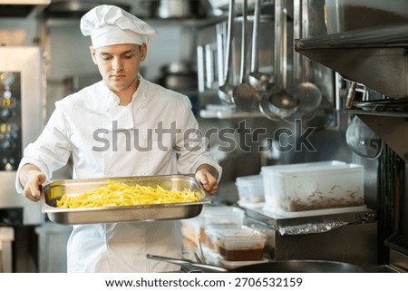 Young man cook in kitchen is at work. Kitchen worker in white uniform performs tasks of chef and auxiliary functions, cooking French fries.