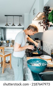 A Young Man Is Cooking Cheese Pancakes In A Modern Light Kitchen At Home. Man Preparing Pancakes For Frying On The Pan. Housework And Home Duties. Culinary Hobby. Vertical Card. Selective Focus