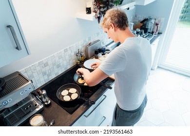 A Young Man Is Cooking Cheese Pancakes In A Modern Light Kitchen At Home. Man Preparing Pancakes For Frying On The Pan. Housework And Home Duties. Culinary Hobby.High View. Selective Focus