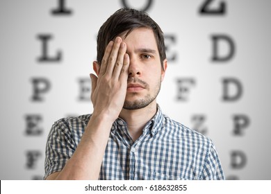 Young Man Is Covering His Face With Hand And Checking His Vision. Chart For Eye Sight Testing In Background.