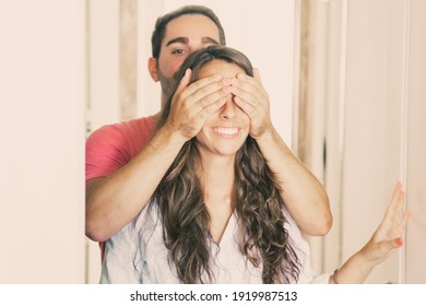 Young Man Covering His Girlfriend Eyes With Hands And Leading Her Into Their New Apartment. Family Couple Walking Through Doorway. Medium Shot. New Home Or Surprise Concept