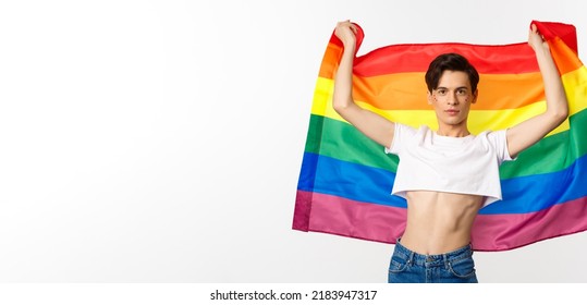 Young Man In Crop Top, With Glitter On Face, Raising Pride Flag With Confident Emotion. Queer Person Smiling With A Lgbt Flag