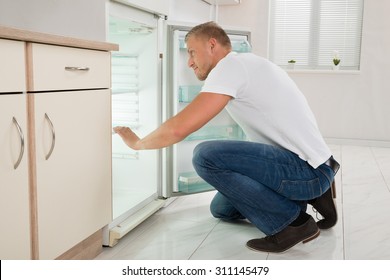 Young Man Crouching On Floor And Looking Into An Empty Refrigerator In Kitchen