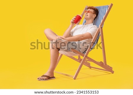 Young man with cup of soda resting in deck chair on yellow background