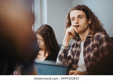 A young man with curly hair appears thoughtful while attending a business meeting. Colleagues in the background focus on their work. - Powered by Shutterstock