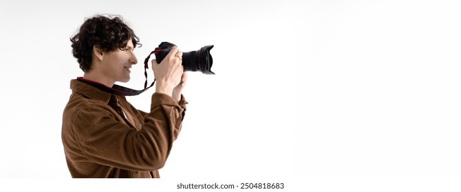 A young man with curly hair is enthusiastically taking pictures using a professional camera in a brightly lit studio. His focus is evident as he aims the lens towards the subject, copy space - Powered by Shutterstock