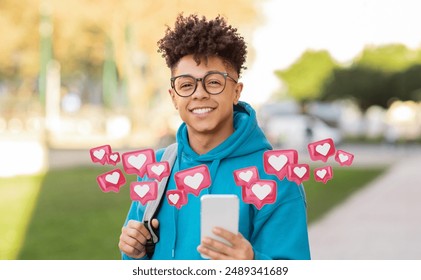 A young man with curly hair and glasses is smiling while holding a smartphone in his hand, and looking at the camera. He is standing outside during the daytime, and there are several red heart icons - Powered by Shutterstock