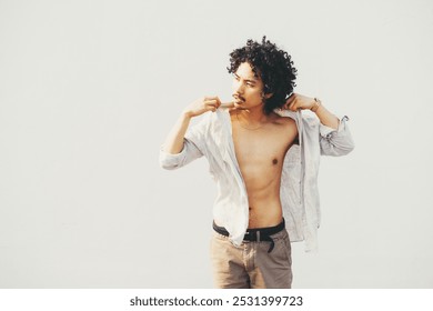 A young man with curly hair poses against a light background, unbuttoning his shirt. He has an intense gaze, wearing beige pants with a black belt, showing a casual fashion style - Powered by Shutterstock