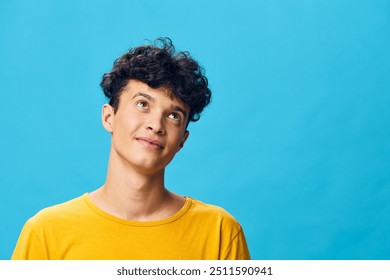 Young man with curly hair wearing a yellow shirt, looking upwards against a bright blue background, conveying curiosity and positivity Ideal for youth oriented concepts - Powered by Shutterstock