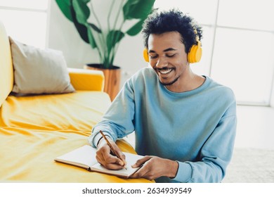 Young man with curly hair wearing yellow headphones and writing in a notebook while sitting in comfortable home interior