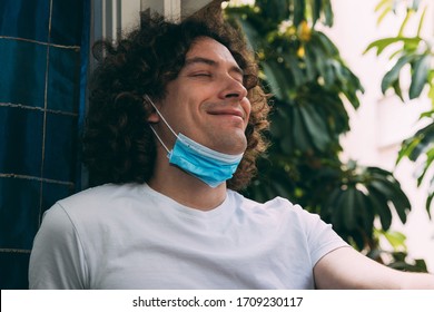 A young man with curly long hair, with a medical mask removed from his face, closed his eyes and smiles. End of home quarantine and the coronavirus epidemic. Good mood. New reality - Powered by Shutterstock