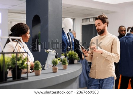 Young man customer using mobile loyalty program app while standing at checkout cashier counter in clothing store. Shopper buying clothes in shopping mall paying with smartphone