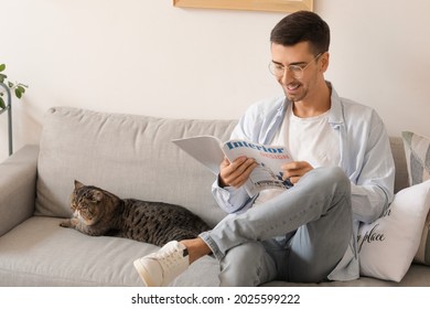 Young Man With Cute Cat Reading Magazine At Home