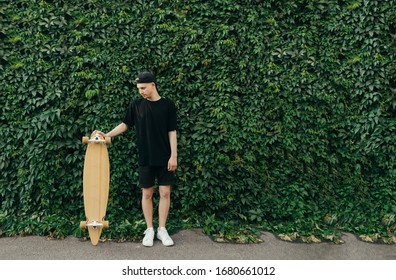 Young man in dark clothing and a cap stands on the background of an ivy wall, holding a longboard in his hand. Guy with a longboard poses on a background of green wall with ivy.Background
