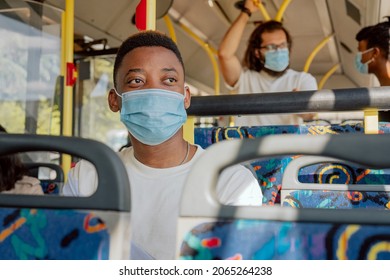 Young Man With Dark Complexion Sits On Public Transport Bus Mask On Face Looking Out Window, Two People In Background Stand Holding Handles And Talking To Each Other Afternoon Return From Work