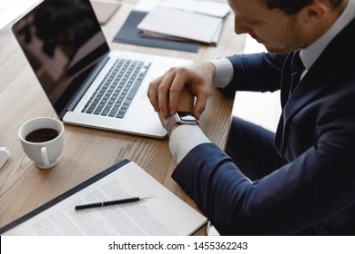 Young man in dark suit sitting at the office table with laptop and documents and looking at the smart watch - Powered by Shutterstock