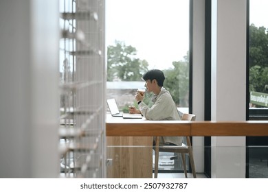 A Young man deeply focused on work in a quiet cafe, sipping coffee while working on a laptop, with a serene outdoor view through large windows. - Powered by Shutterstock